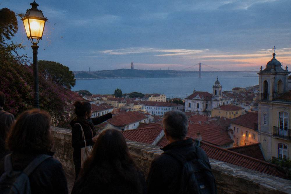A guide gestures to a small group at a Lisbon viewpoint above red rooftops and the Tagus at dusk.