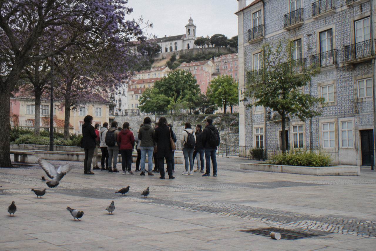A walking tour group stands in a Lisbon square beside azulejo-tiled buildings, with pigeons on the pale stone paving and a hilltop church behind.