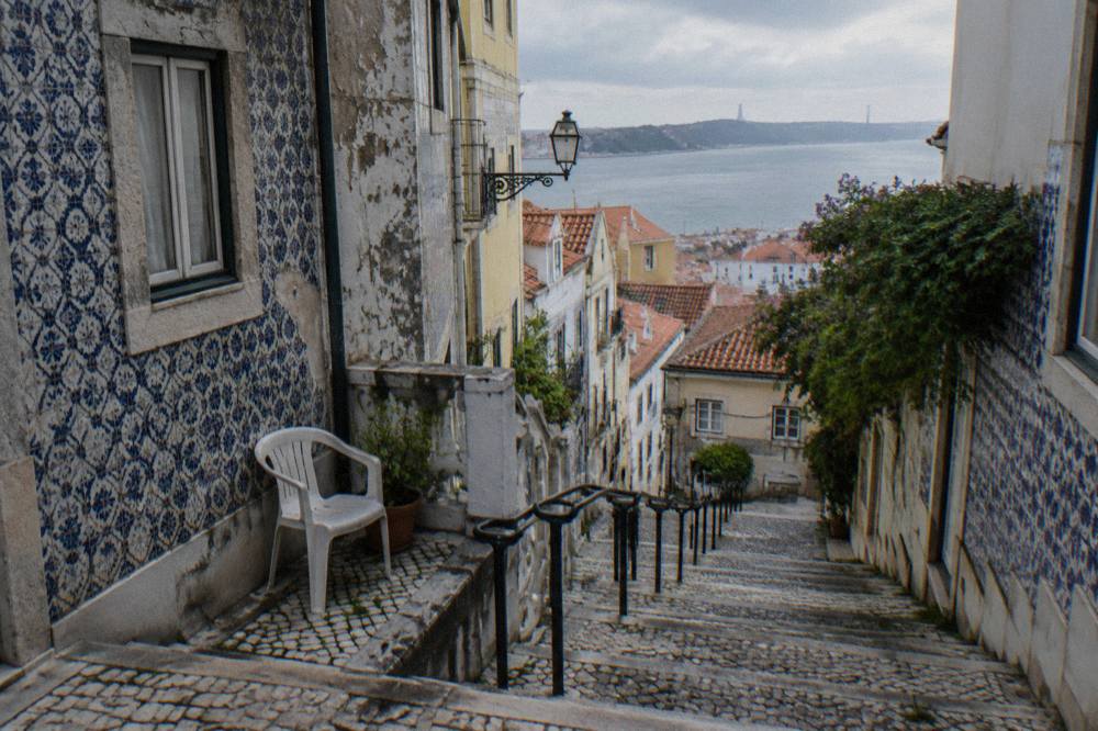 Steep cobbled steps descend between azulejo-tiled houses with a plastic chair, handrails, and a view over rooftops to the Tagus.