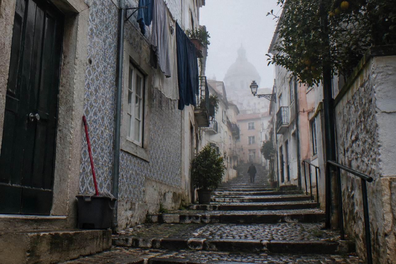 A narrow cobbled uphill street in fog with tiled houses, laundry, a mop bucket, and a distant figure.