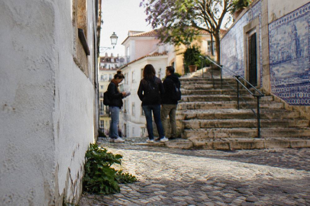 Three people stand on a narrow cobbled Lisbon street beside stone steps and blue azulejo-tiled walls.