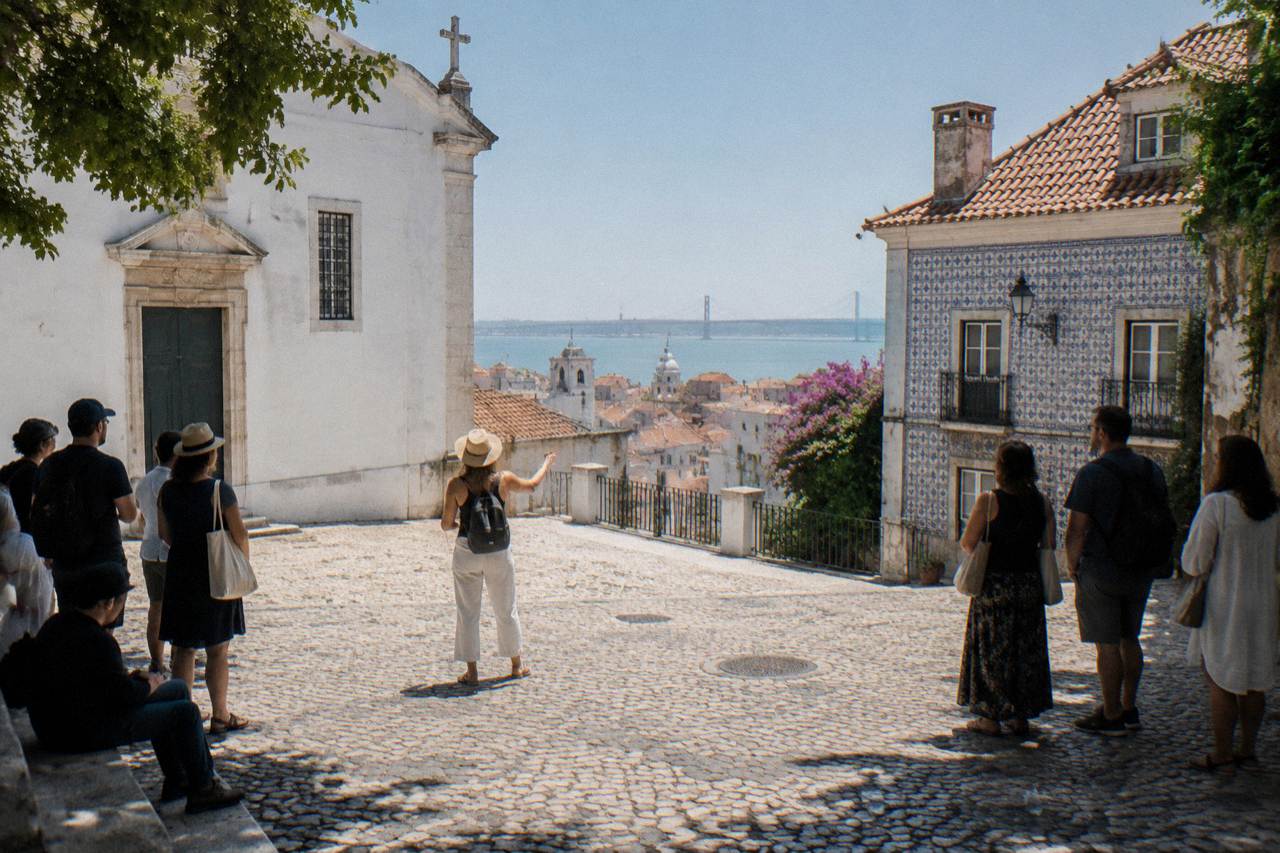 A guide stands in a cobbled Lisbon square facing a small group between a white church and an azulejo-tiled house, with the Tagus beyond.