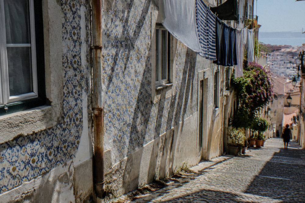 A steep cobbled Lisbon street with a tiled facade, laundry hanging overhead, potted plants, and a distant figure descending.