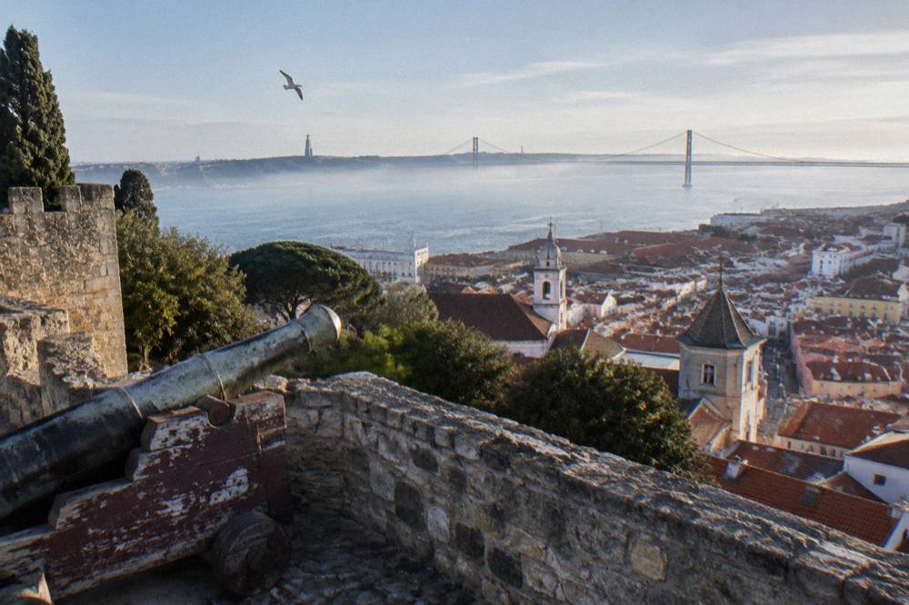 Stone castle ramparts overlook Lisbon rooftops, church towers, the Tagus estuary, a bridge, and a gull in the sky.