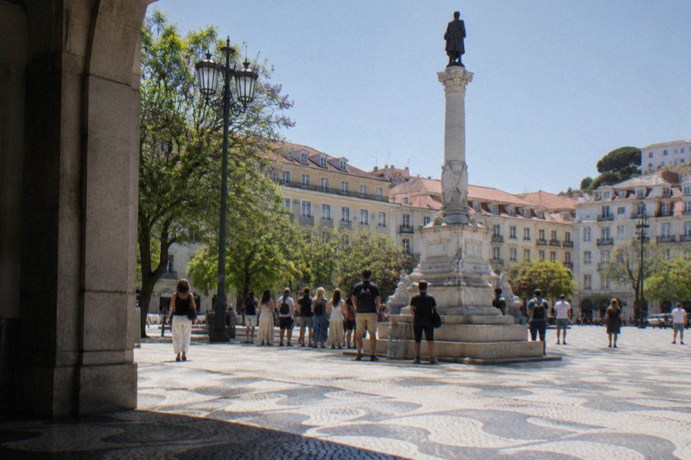 People stand around a tall column monument in a sunlit Lisbon square with patterned cobblestones and surrounding apartment facades.