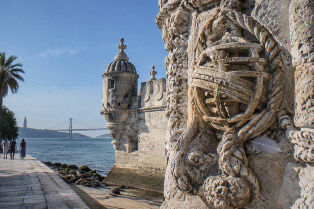 Close view of Belém Tower’s carved stone facade beside the riverside walkway with the Tagus and bridge in the background.