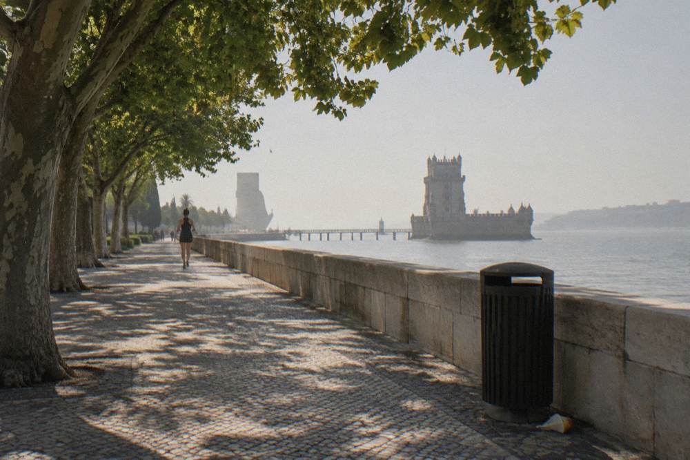 Tree-shaded riverside promenade with a distant stone tower across the water and a lone jogger on the path.