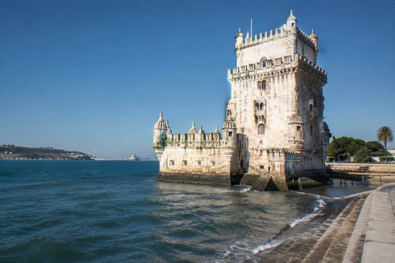 Belém Tower stands beside the Tagus with stone steps in the foreground and a clear blue sky behind.