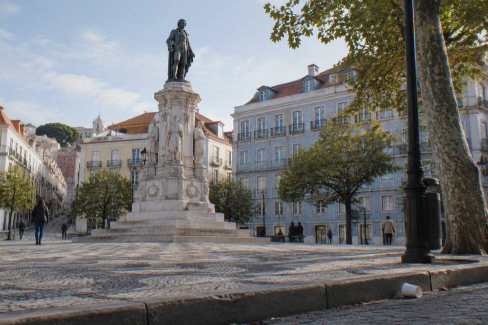 A bronze statue stands on a white stone pedestal in a cobbled Lisbon square with pale tiled facades, trees, and a dropped cup by the curb.