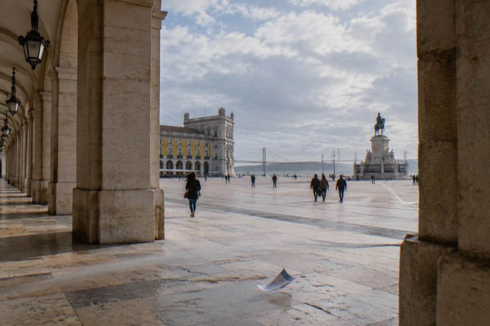 Arcaded limestone colonnade overlooks Praça do Comércio with a wide square, distant statue, a few pedestrians, and a flyer on the pavement.