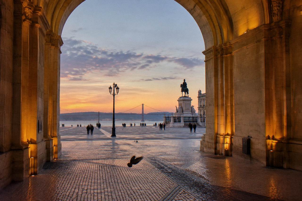 View through a stone arch onto Praça do Comércio at sunset with a lamppost, equestrian statue, distant bridge, and a pigeon on cobbles.
