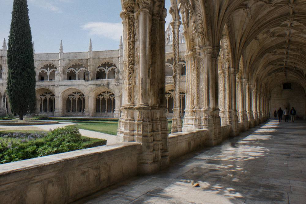 Stone cloister arcade with carved arches around a courtyard, sunlight on the flagstone walkway, and a few distant people.