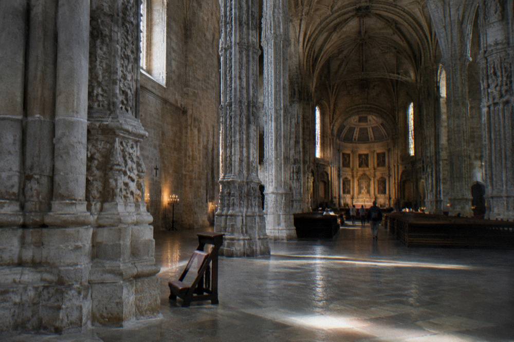 A vast stone church interior with tall columns, ribbed vaults, side windows, and a small kneeler on the floor.