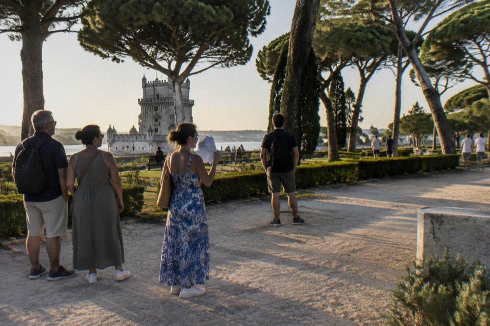 Three visitors stand on a gravel path in a riverside garden with trees and the Belém Tower beyond.