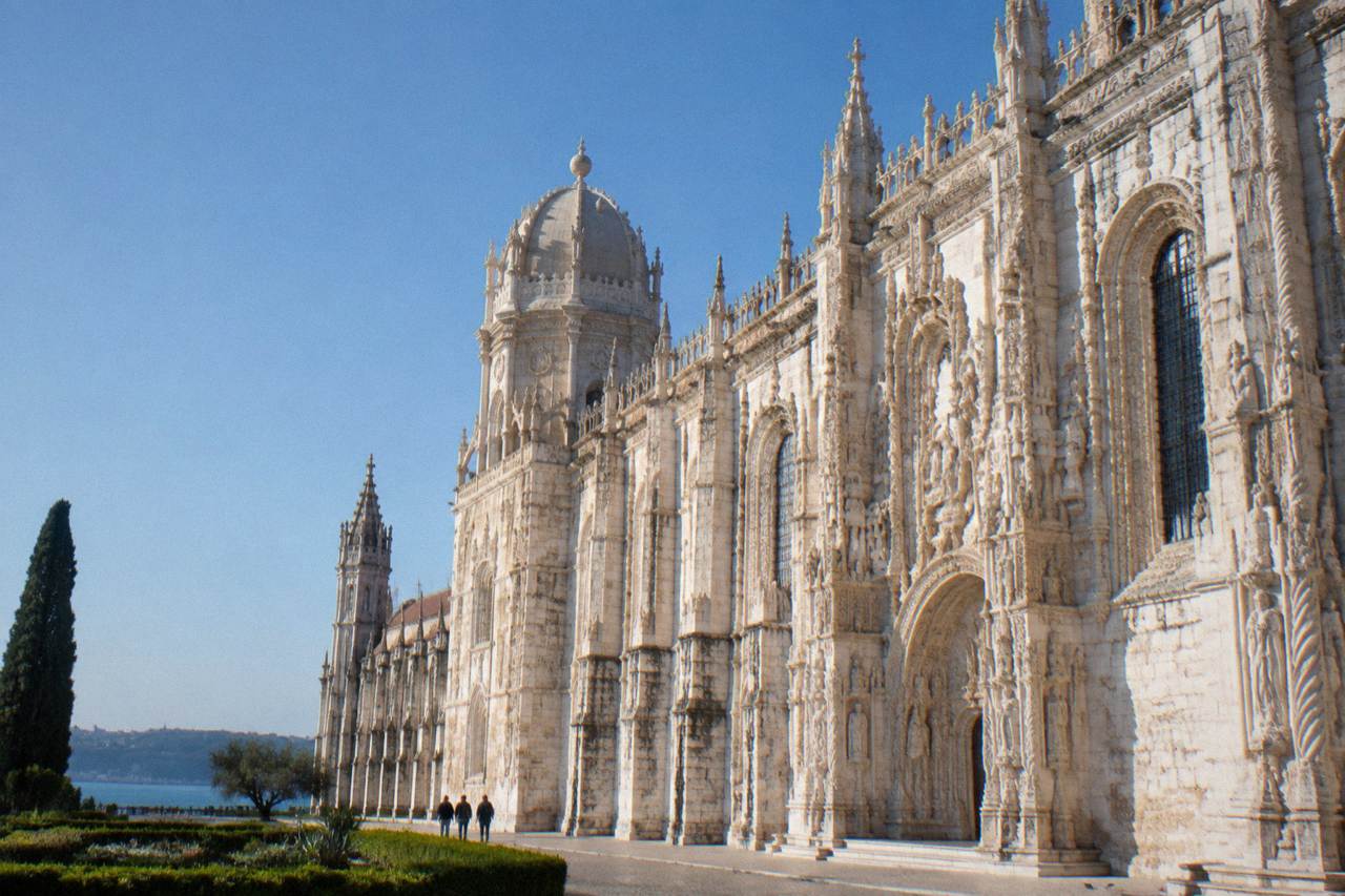 The stone facade of Jeronimos Monastery rises under a clear blue sky beside a formal garden and distant water.