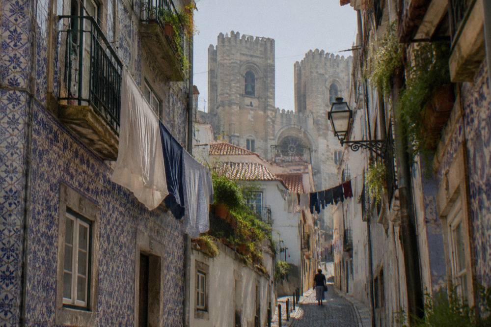 A narrow cobbled lane with hanging laundry leads toward Lisbon Cathedral bell towers above tiled houses.