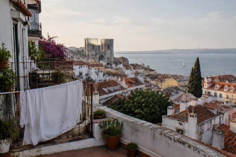 Terrace with a drying sheet overlooks Alfama rooftops, Lisbon Cathedral, and the Tagus under a pale morning sky.
