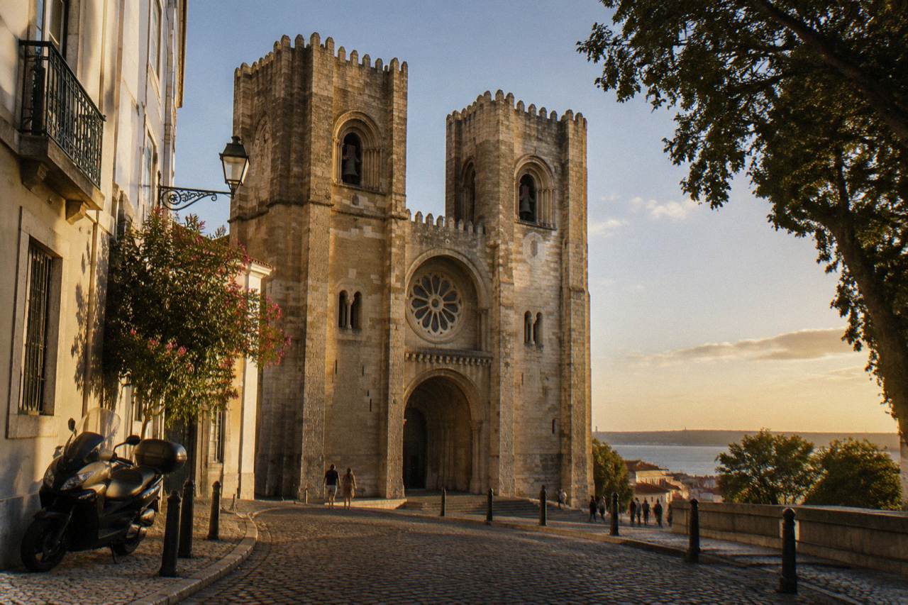 Lisbon Cathedral rises over a cobblestone square at sunset, with a parked scooter at left and the estuary visible beyond.