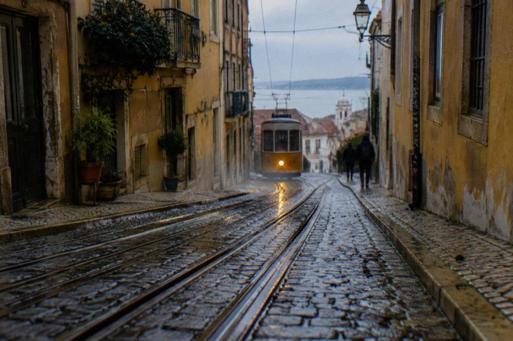 A wet steep Lisbon street with tram tracks, old yellow buildings, a distant tram, and the river beyond.