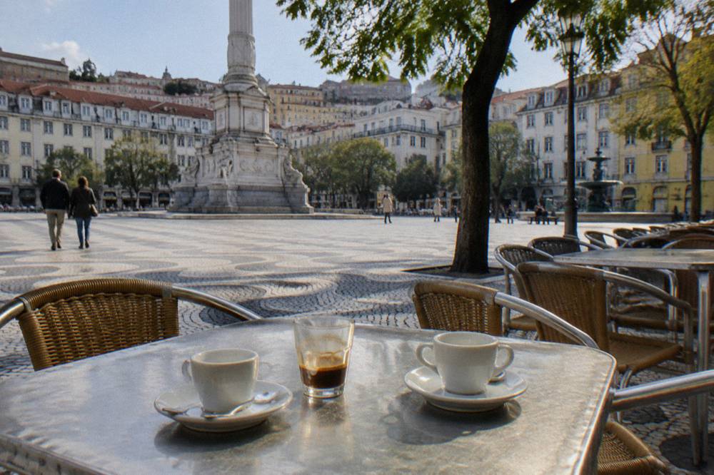 Two empty coffee cups and a small glass sit on a metal café table beside a tiled Lisbon square with a monument and hillside buildings.