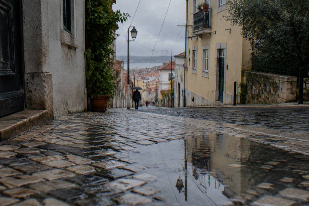 A wet cobblestone street slopes downhill between pale buildings with a puddle reflecting a lamp post and the city beyond.