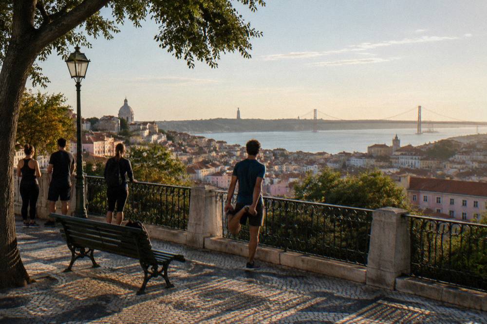 Four people stand and stretch on a tiled Lisbon viewpoint above rooftops and the river with a bridge in the distance.