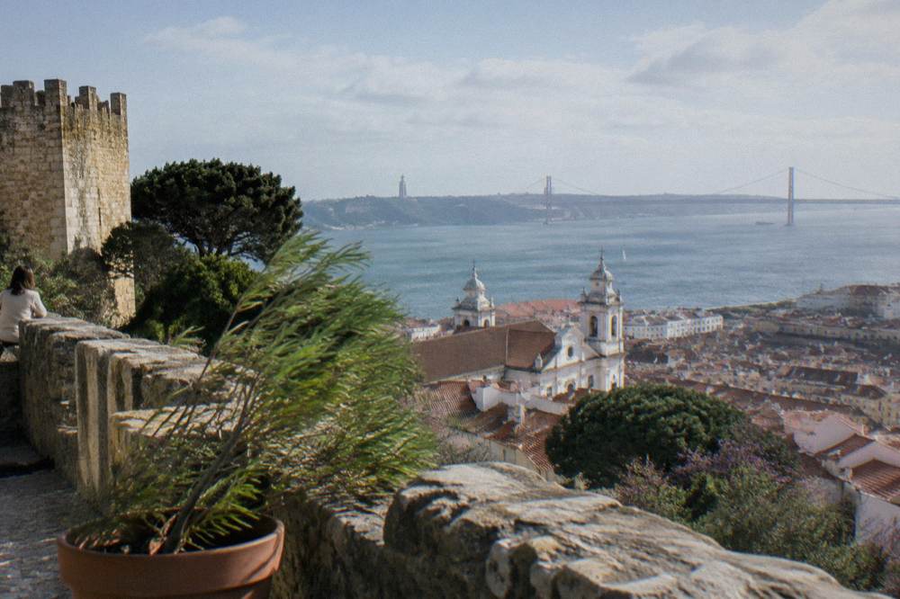Stone castle wall and terrace overlook Lisbon rooftops, church towers, the Tagus River, and the 25 de Abril Bridge.