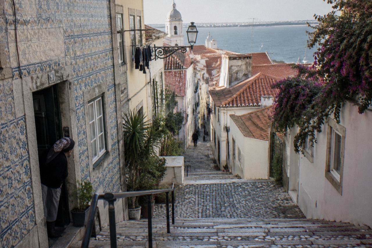 A steep cobbled Lisbon lane descends between tiled houses with a woman leaning from a doorway and the river beyond.