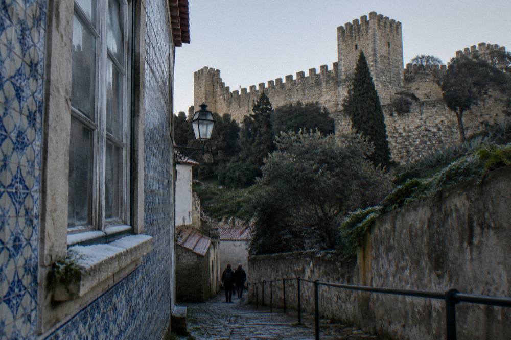 A narrow tiled lane climbs toward crenellated castle walls above trees, with a blue azulejo facade and a few small figures below.