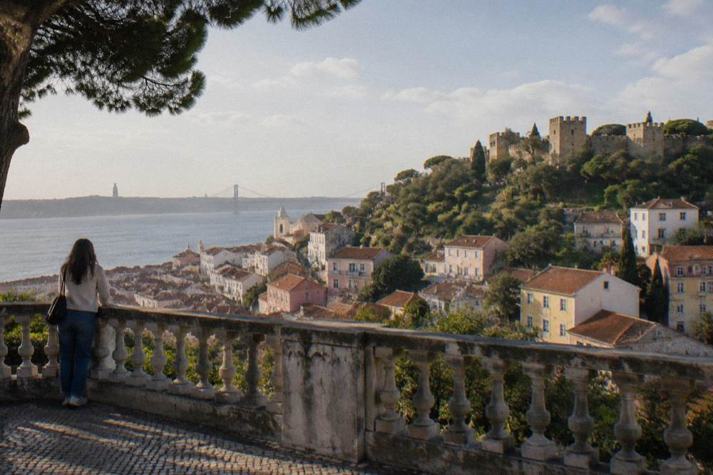 A woman stands by a stone balustrade overlooking Lisbon rooftops and São Jorge Castle across the Tagus.