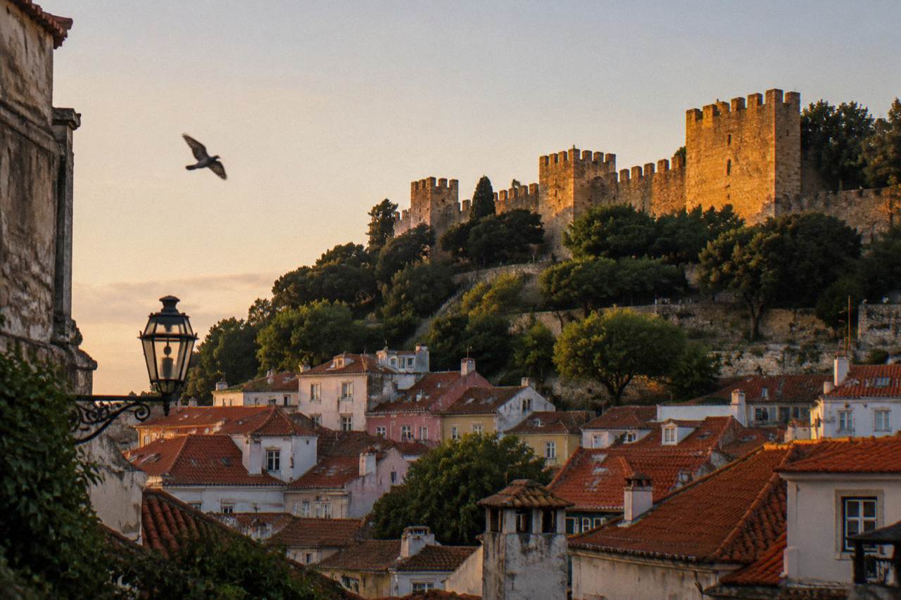 Stone castle walls and towers glow above red-tiled rooftops, with a bird in the sky and a lamp on the left.