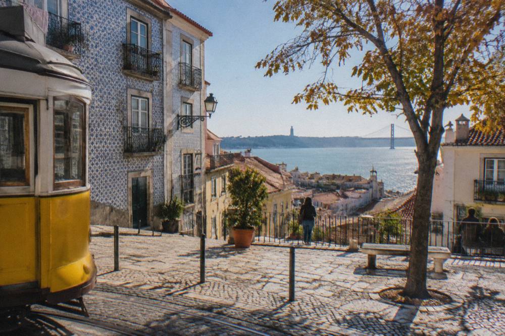 A cobbled Lisbon square with a yellow tram, tiled facades, a tree, and a view over rooftops to the river and bridge.
