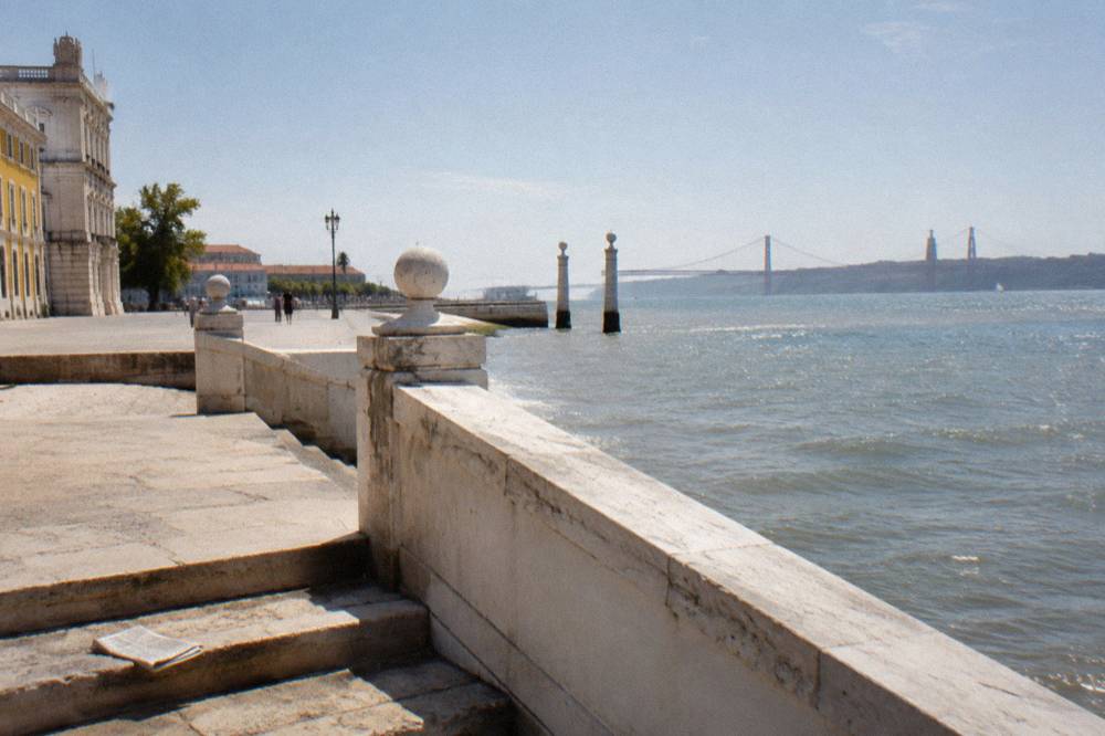 Stone balustrade steps beside a broad riverfront terrace with open water and a distant suspension bridge.