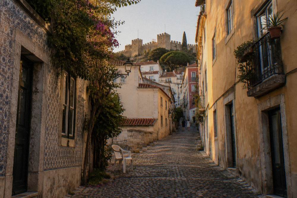 A steep cobbled Lisbon street climbs between tiled old buildings toward castle walls, with a white plastic chair in the foreground.