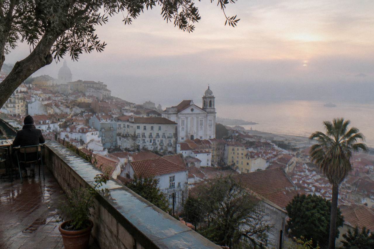 A misty dawn view from a terrace over Lisbon rooftops toward the Tagus with a seated person, church towers, and a palm tree.