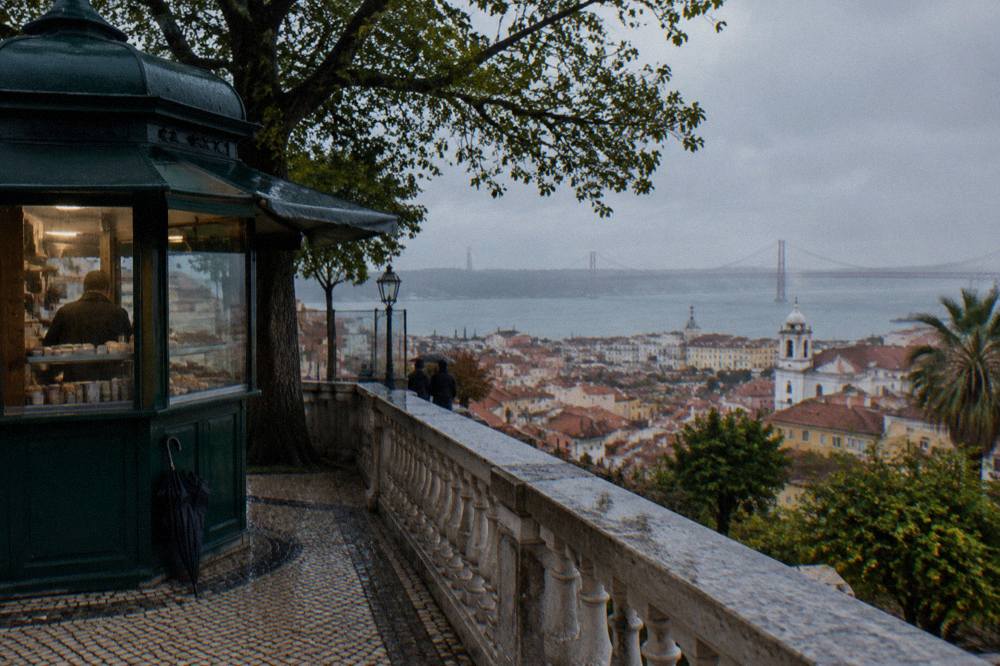 A stone terrace with a green kiosk overlooks Lisbon rooftops and the river on a gray rainy day.