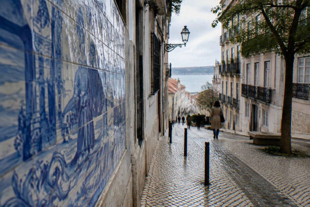 Blue and white azulejo panels line a narrow sloping cobbled street with a few pedestrians, balconies, a tree, and water in the distance.