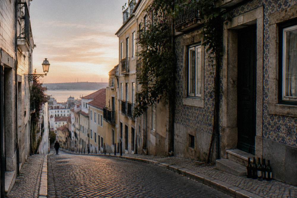 An empty steep cobbled street lined with tiled buildings and balconies descends toward the river at sunrise.