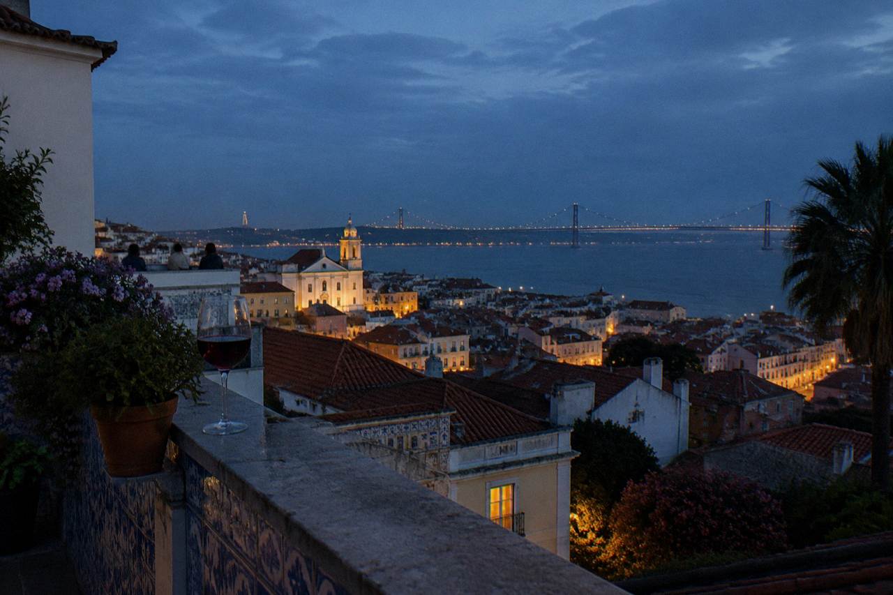 A tiled terrace with a wine glass overlooks Lisbon rooftops and the illuminated Tagus at dusk.