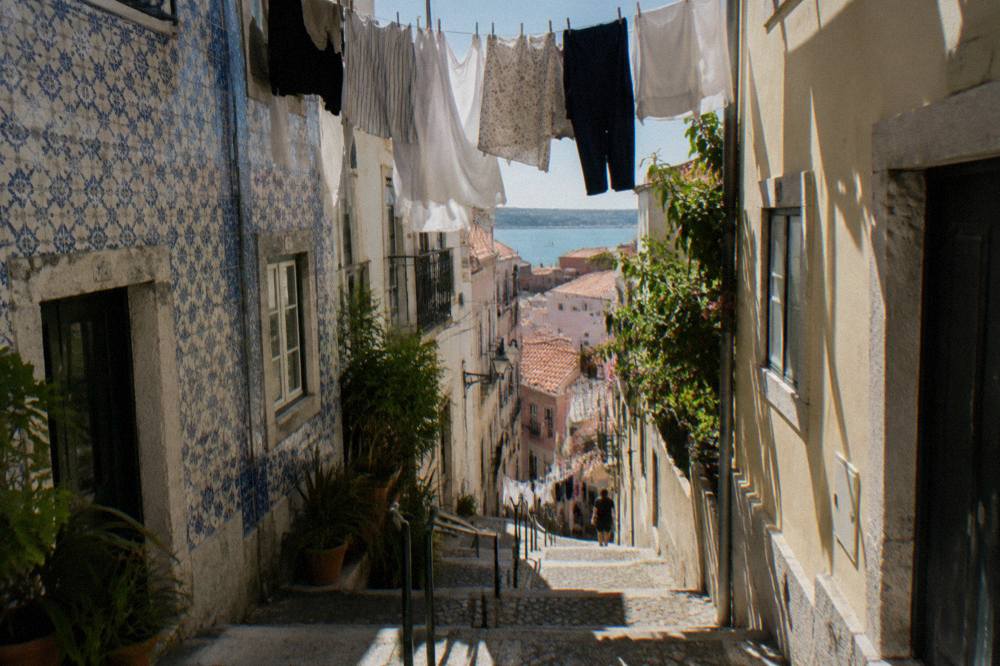 A steep narrow Lisbon lane with worn steps, tiled facades, laundry overhead, and a distant view toward the water.