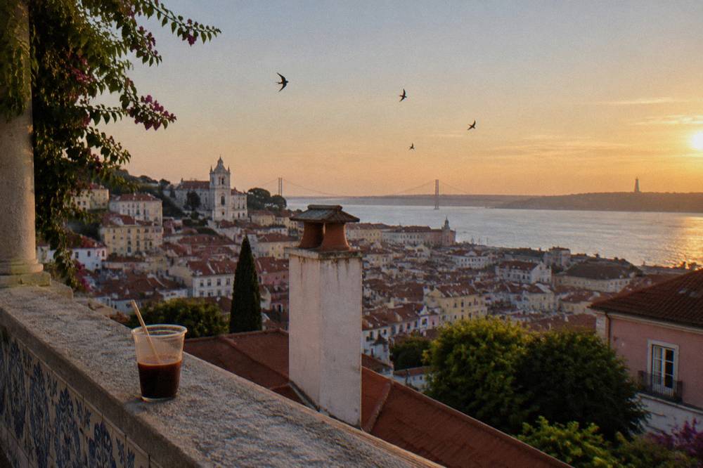 A tiled terrace overlooks Lisbon rooftops, the Tagus river, a bridge, and birds in warm sunset light, with a coffee cup on the parapet.