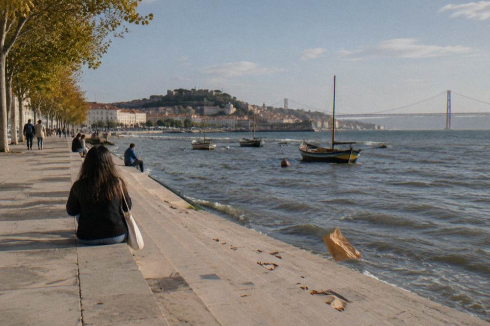 Stone riverside steps with seated people, moored boats on the Tagus, and a distant Lisbon skyline with bridge in view.