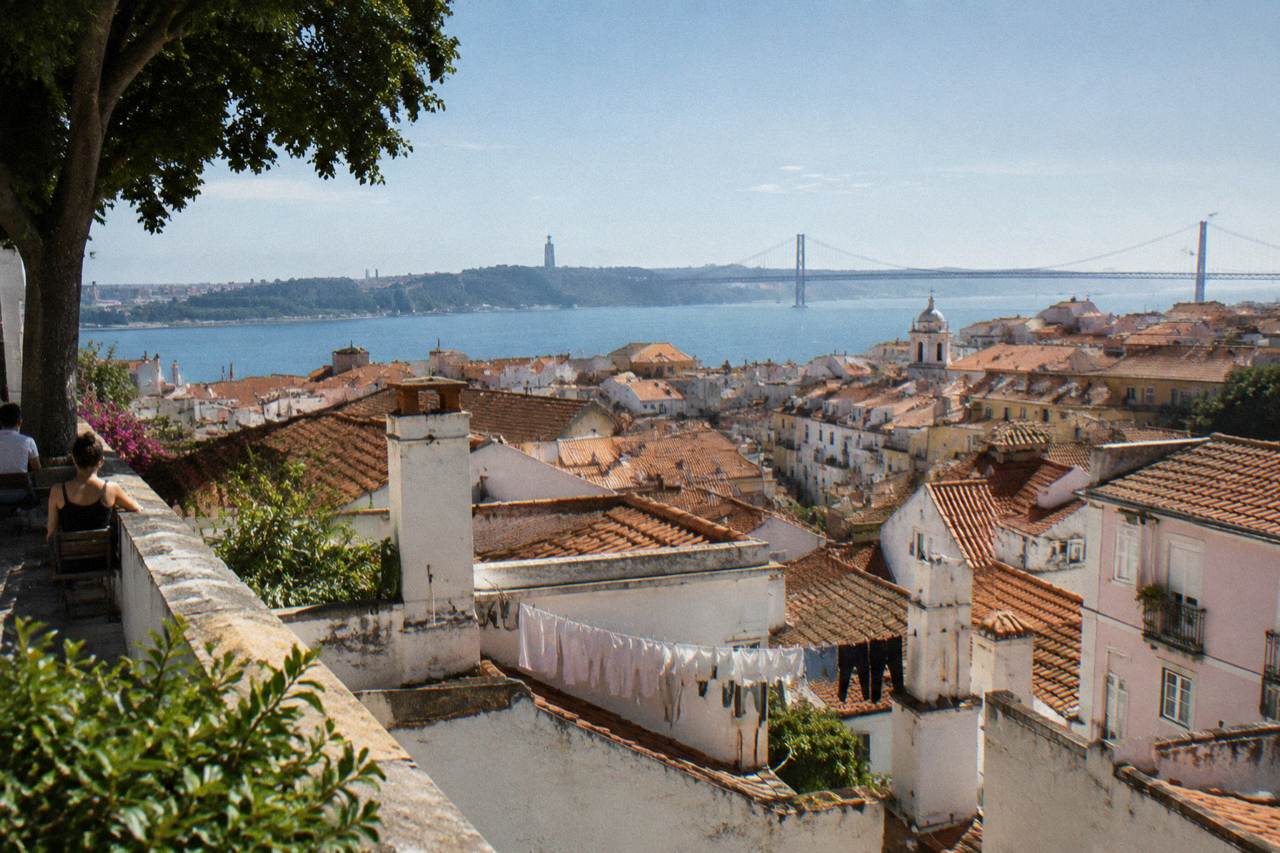 A hilltop terrace looks over Alfama rooftops toward the Tagus River and a distant bridge, with laundry strung between chimneys.