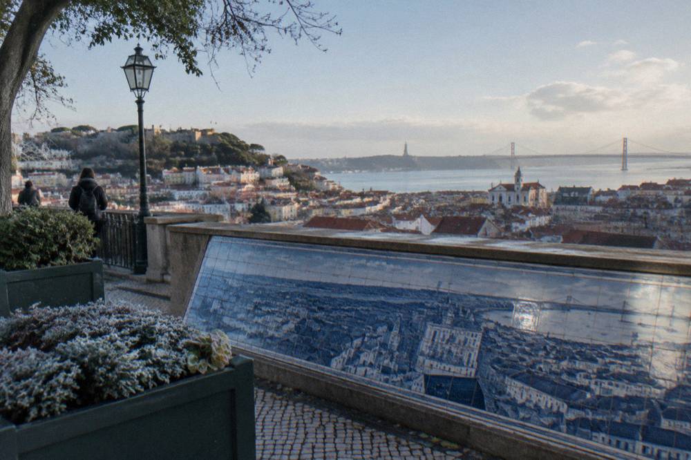 A Lisbon viewpoint terrace with a tiled city map, frosty planters, benches and a wide river and rooftops view.