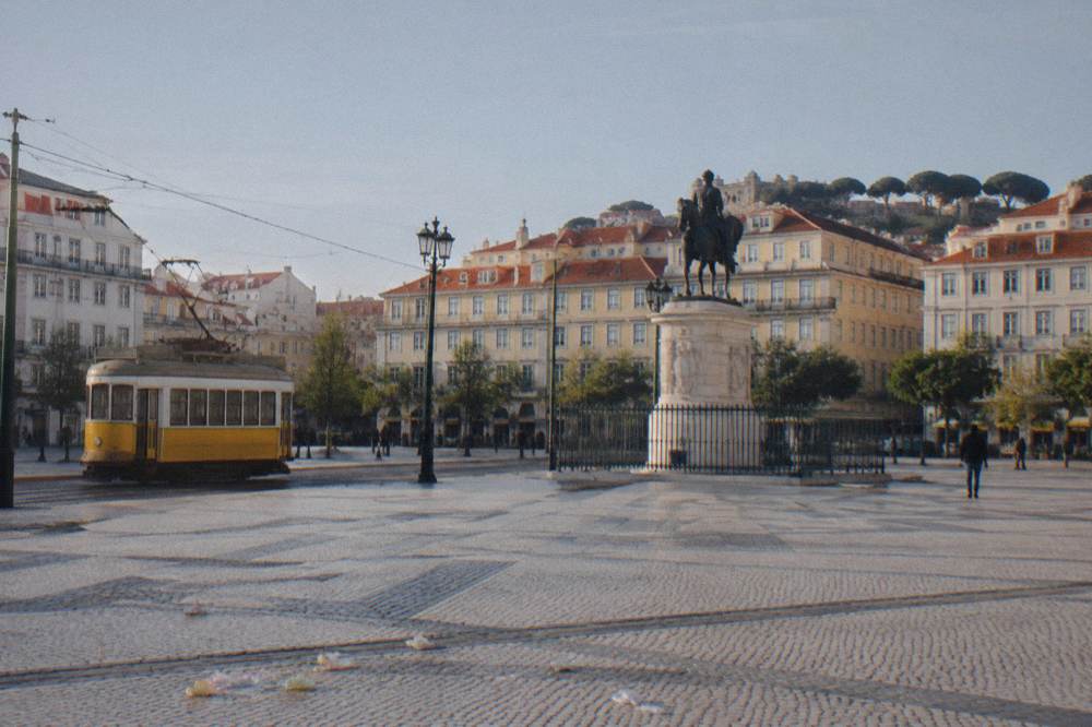 A wide cobbled Lisbon square with a yellow tram on tracks and an equestrian statue on a pedestal.