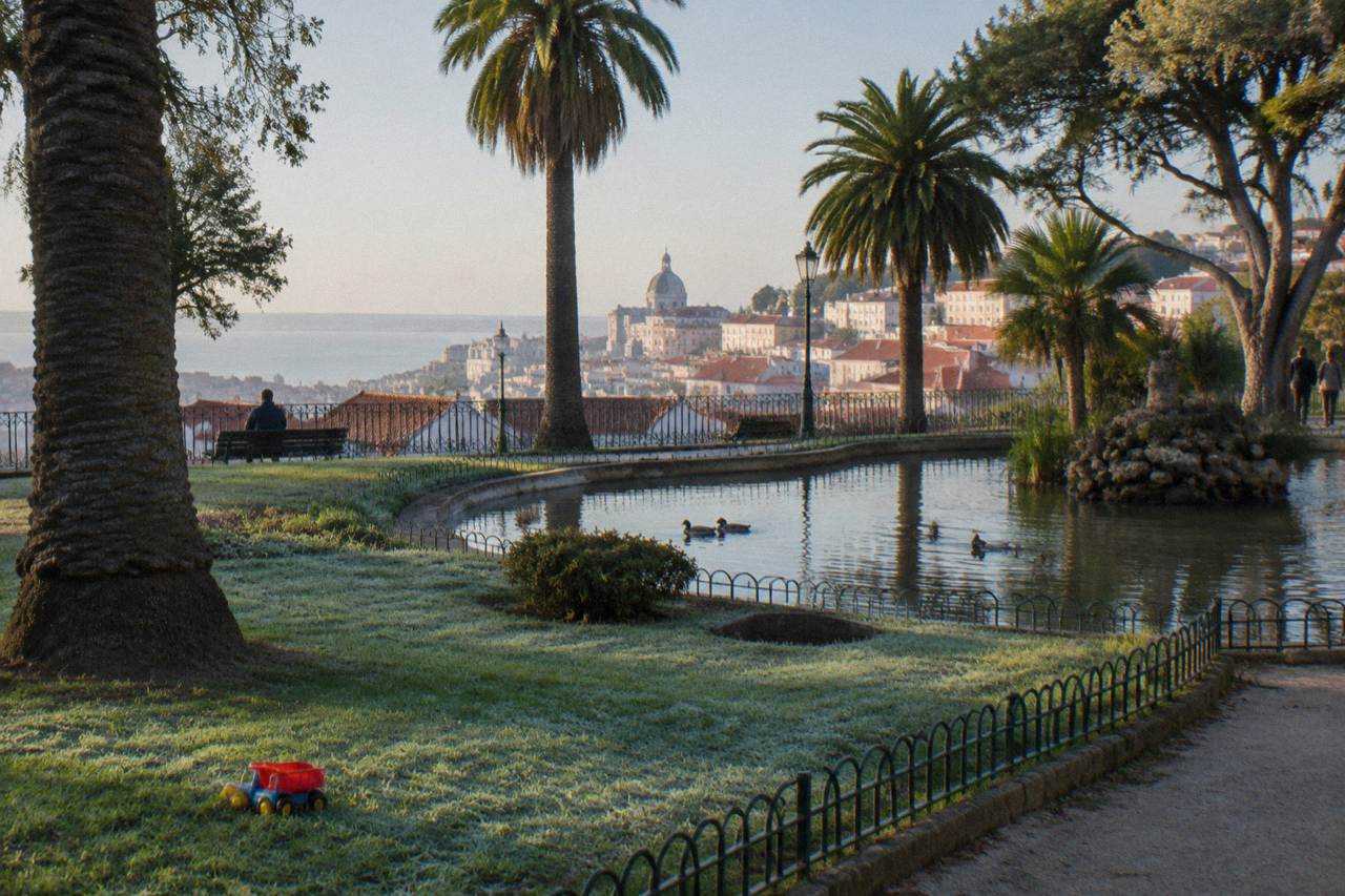 A frosty garden with palm trees, a pond with ducks, a toy truck on grass, and Lisbon rooftops and a river view beyond the railings.