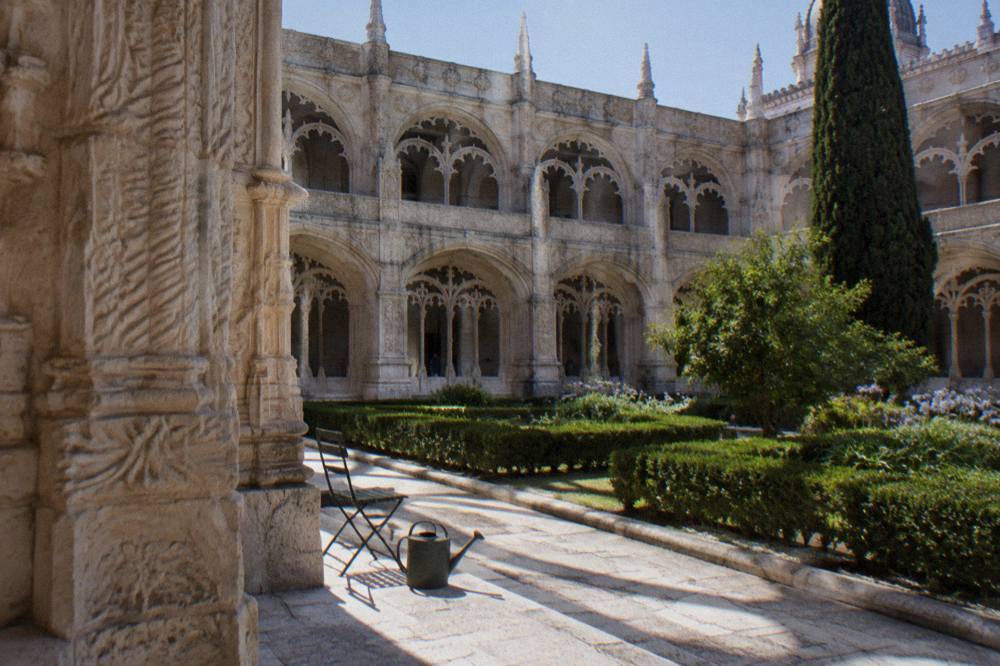 A stone monastery cloister courtyard with carved arches, a folding chair, a watering can, and trimmed hedges in bright sun.