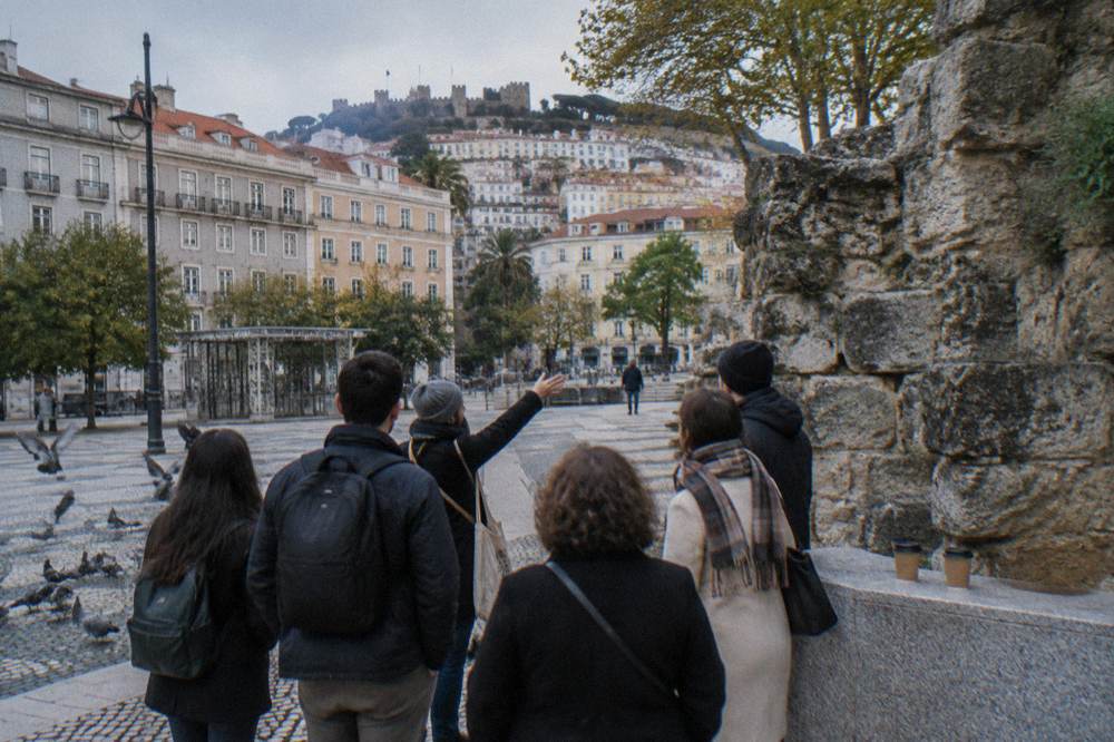 A small group stands by a rough stone wall in a Lisbon square with pigeons on the paving and a castle on the hill beyond.