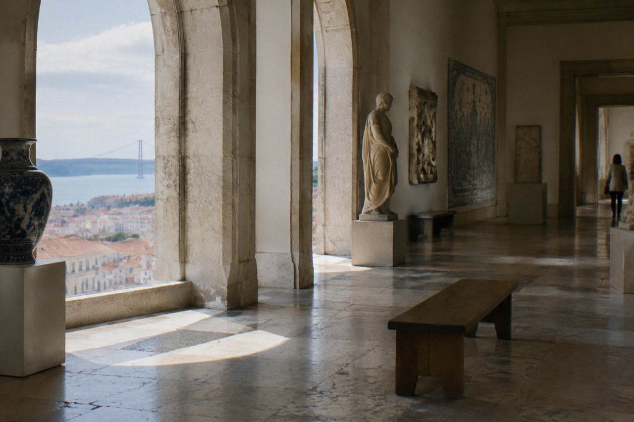 A stone-arched museum gallery with a wooden bench, sculptures, tiled wall panels, and daylight over Lisbon and the river beyond.