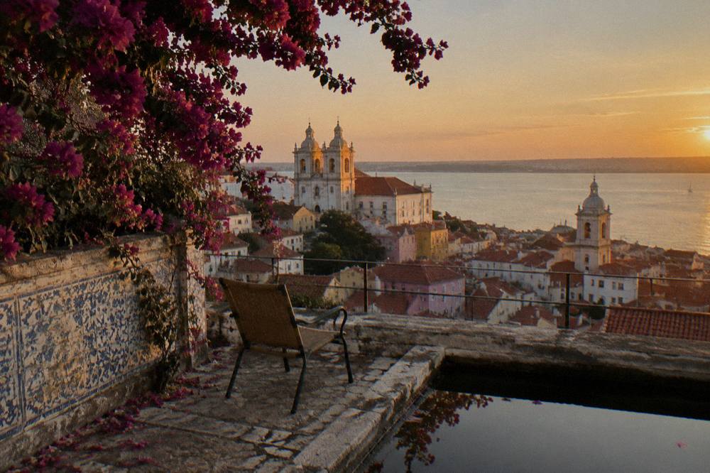 A tiled terrace with a chair, bougainvillea, rooftop Lisbon skyline, church towers, and the river at sunset.